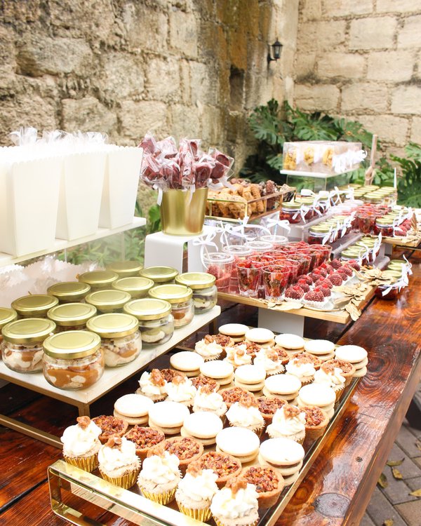 Mesa de postres al aire libre con muro de piedra y bases doradas en evento de jardín